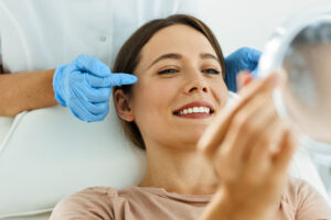 Woman smiling while examining her reflection in a mirror, with a healthcare professional pointing at her face, showcasing aesthetic treatment at Galen Healthcare & Aesthetics.